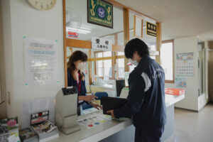 明るく清潔感のある教習所の受付窓口で、親切な日本人指導員が学生に料金プランの説明をしている様子。