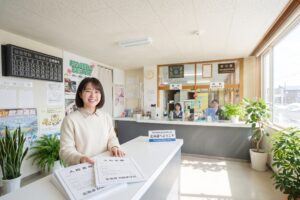北海道の明るく清潔感のある自動車学校の受付カウンターで、入校書類を持って笑顔を見せるアジア人の学生。